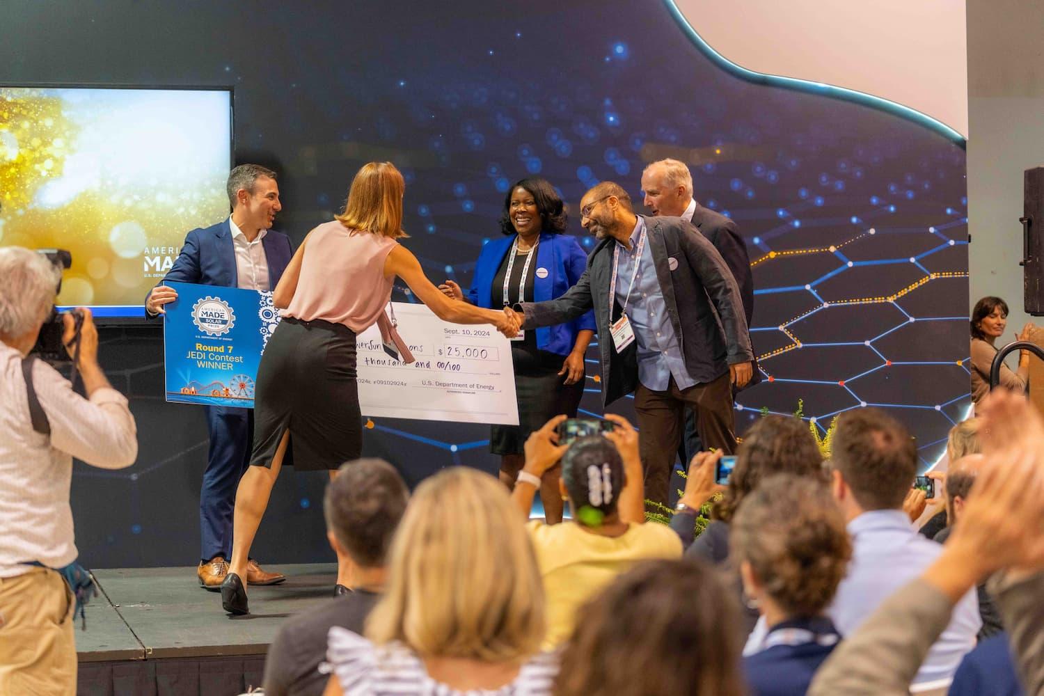 Five people on a stage with an audience in front; two people on stage are handing over a large check and shaking hands with the three people on stage accepting the large check