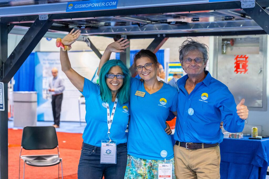 Three smiling people wave and give thumbs up while standing under a large solar panel in a conference hall.