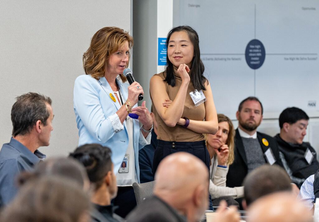 Two people standing up in a room of people, one holding a microphone and talking to the crowd.