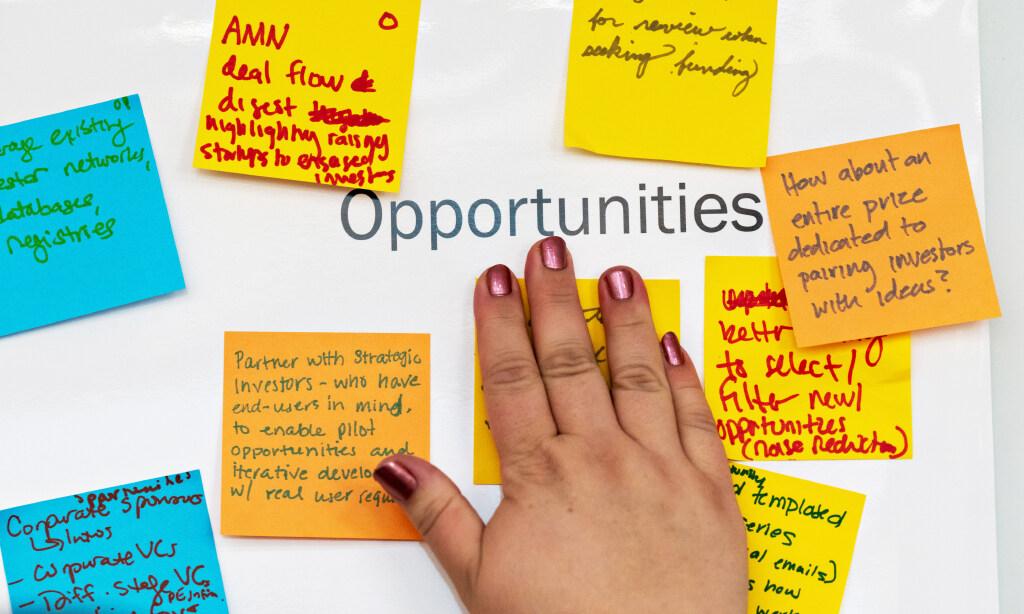 A close-up of a poster that says “opportunities” with colored sticky notes all around and a person’s hand putting up a sticky note